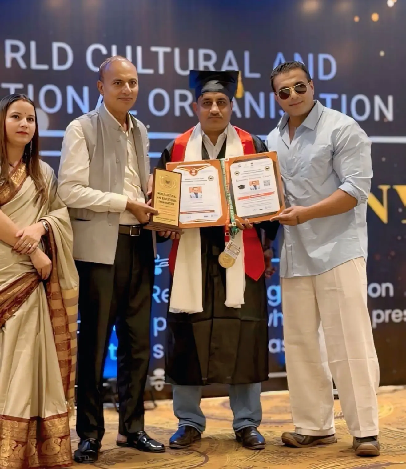 A man in a graduation gown and cap receiving a certificate and plaque on stage at an award ceremony