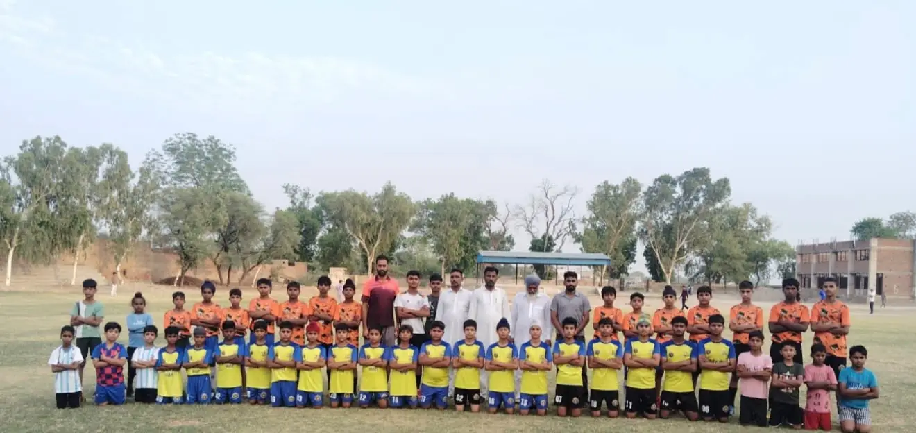 A boys' football team in yellow and orange kits lined up on a ground with their coaches and elders