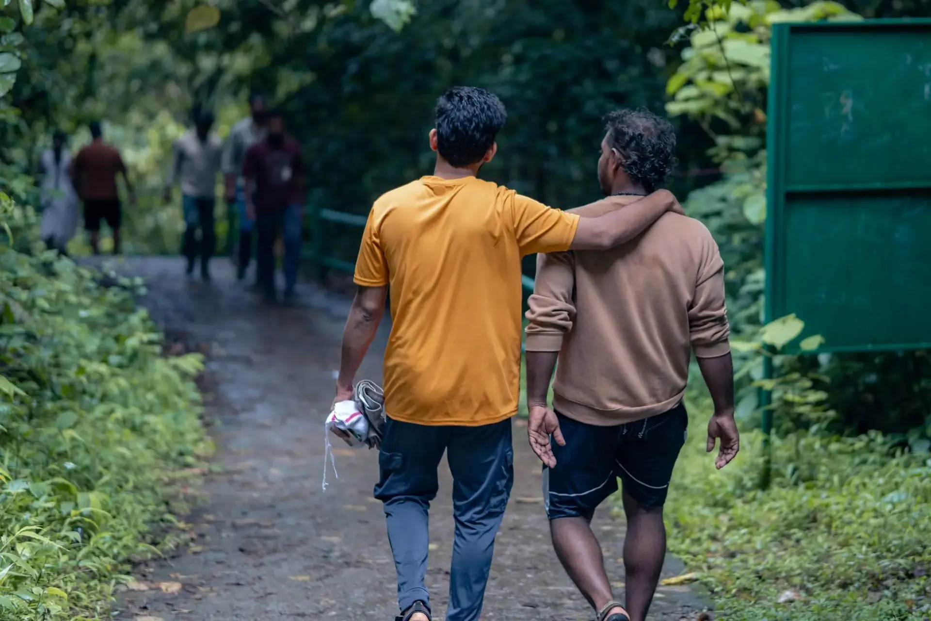 Two men walking a forest path, one with his arm around the other's shoulder